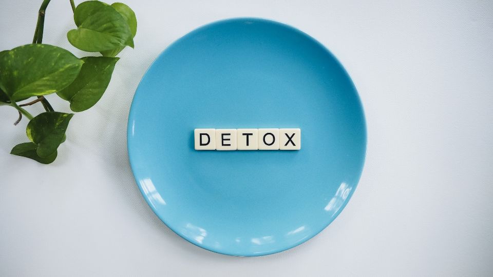Flat lay of a blue plate with the word “DETOX” spelled out in scrabble-style tiles, placed on a white surface next to a green plant, representing a clean and simple detox theme. 